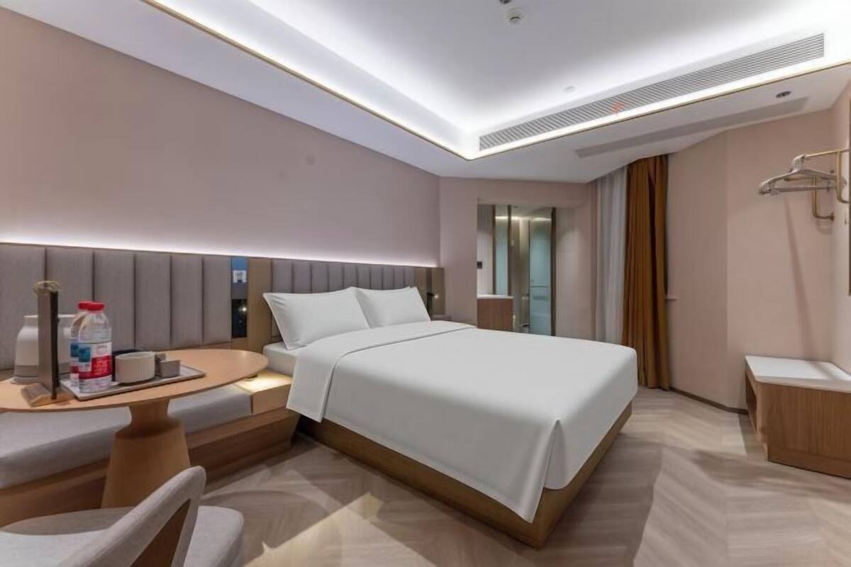 A modern hotel room with a bed, bedside table, chair, and a small desk with a lamp.