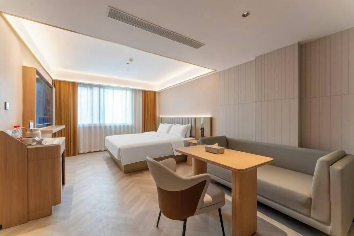 A modern hotel room with a bed, a desk, a chair, a sofa, and a large window with curtains.