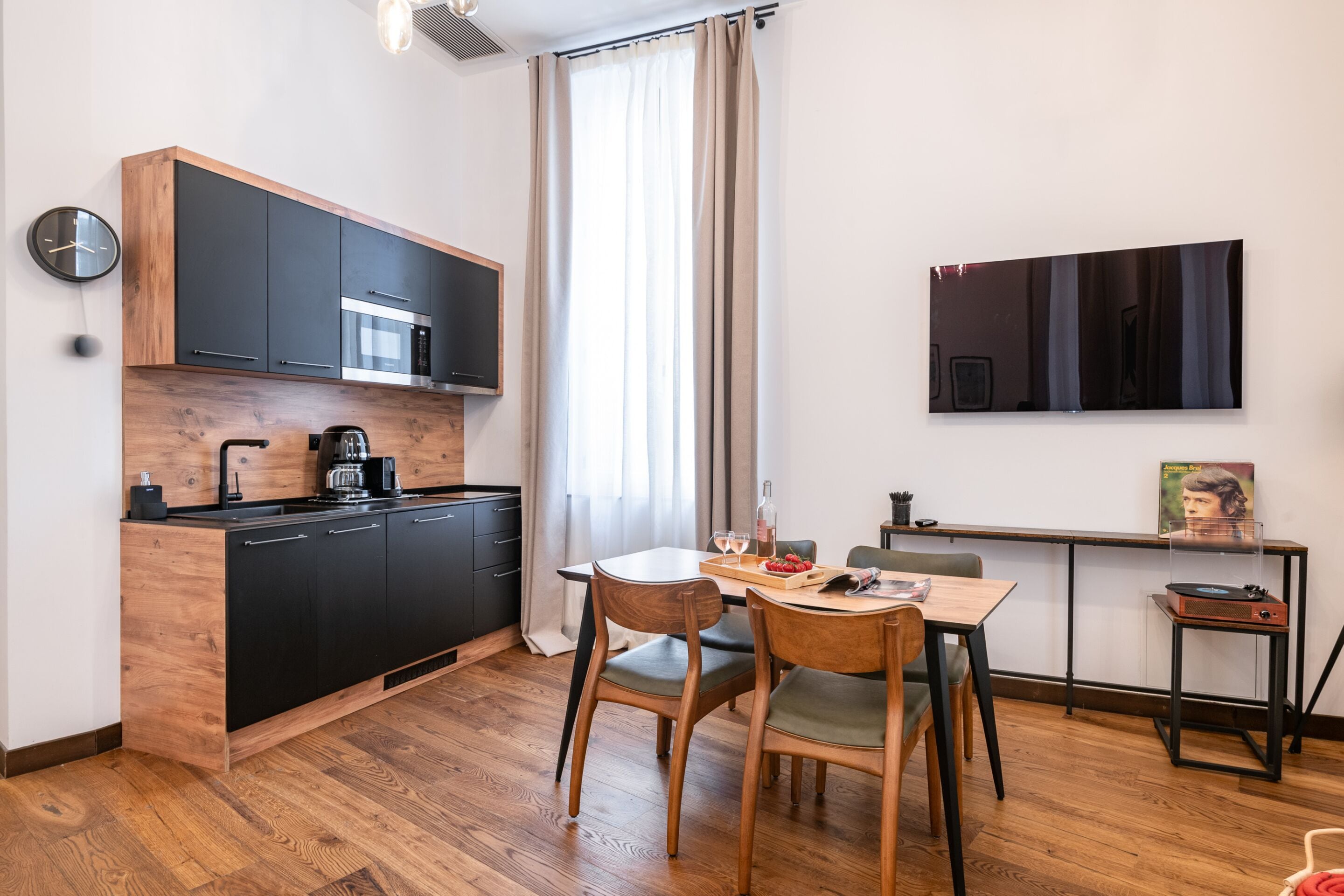 Una cocina moderna con armarios negros, una encimera de madera y una cafetera. Hay una mesa de comedor con sillas y una mesita auxiliar con un tocadiscos.