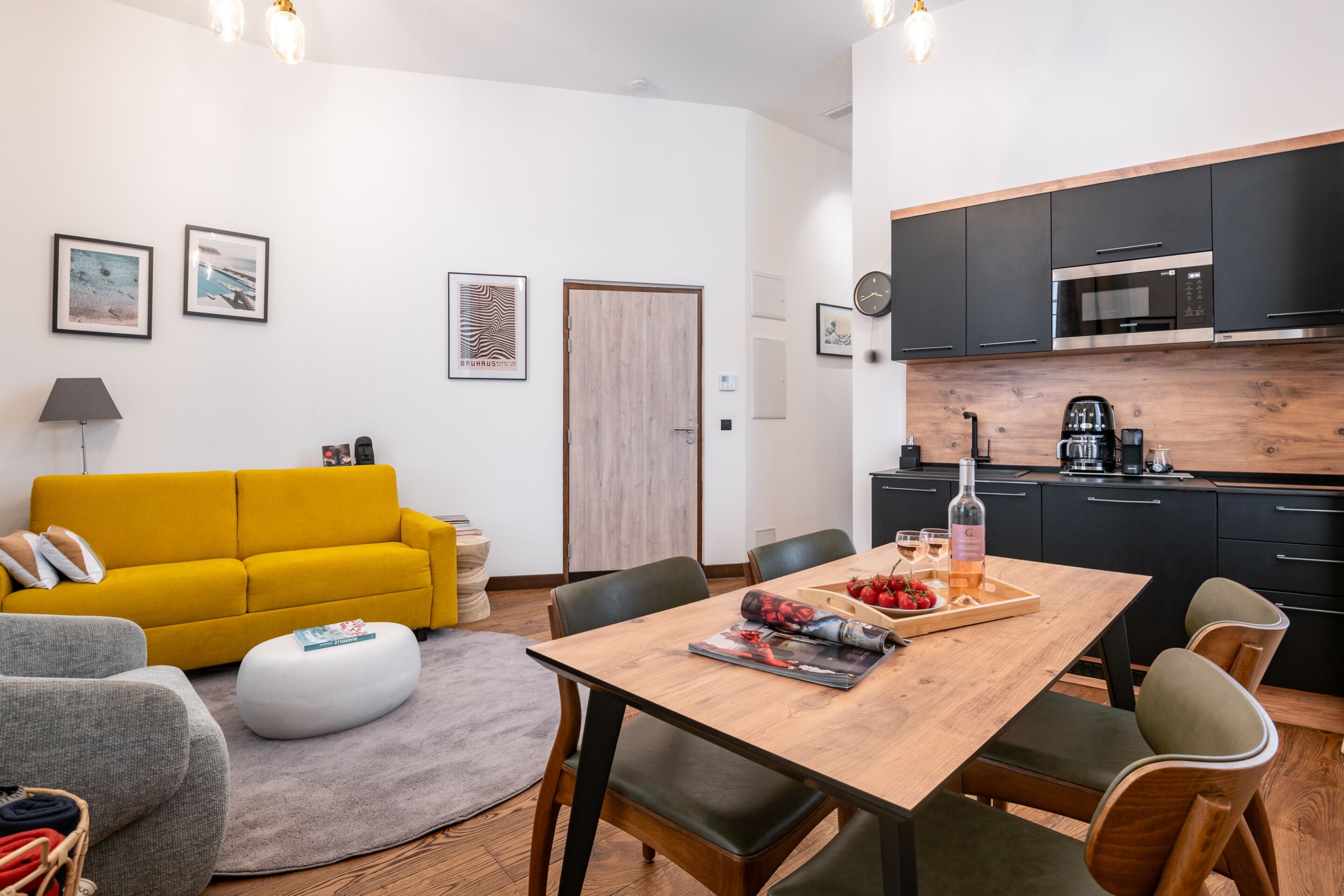 Una sala de estar moderna con un sofá amarillo, una mesa de comedor de madera con sillas, y una cocina con armarios negros y un microondas.