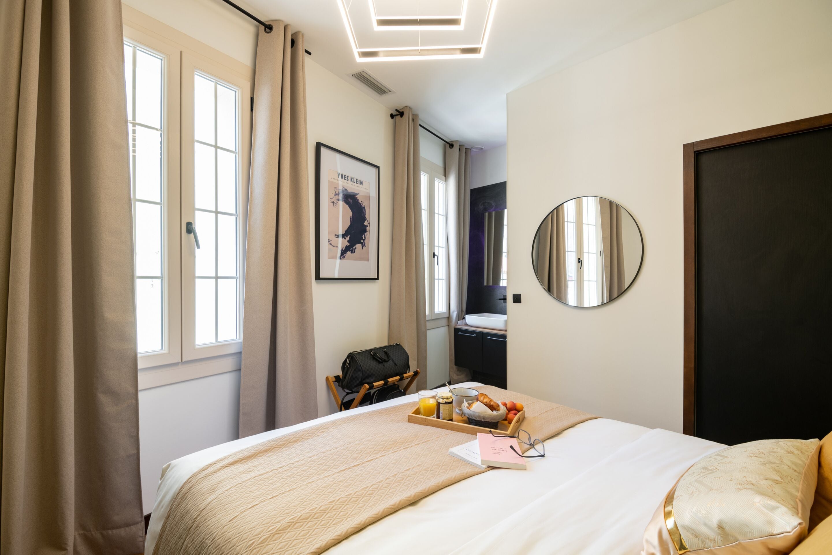 Habitación de hotel con cama, bandeja de desayuno, un cuadro enmarcado, espejo y ventana con cortinas.