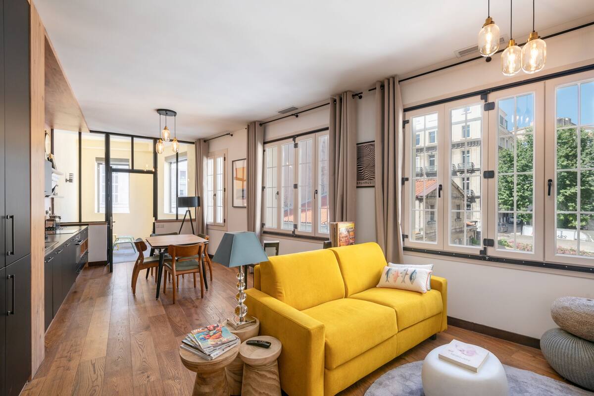A modern living room with a yellow sofa, wooden flooring, and large windows offering a city view.