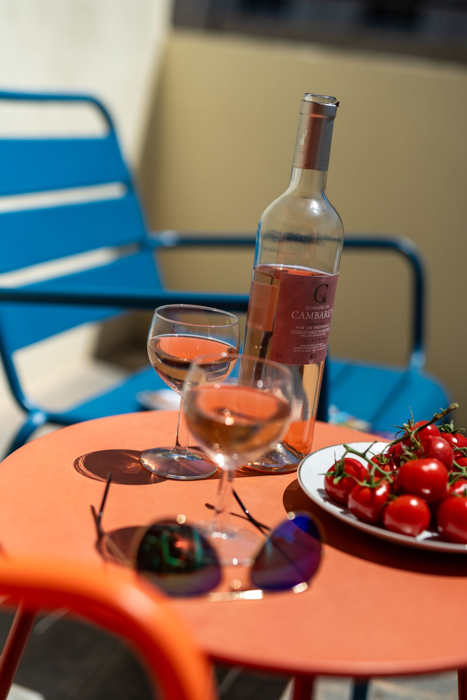 A table with a bottle of wine, two glasses, a plate of cherry tomatoes, and a pair of sunglasses.