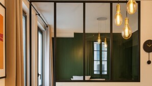 Ein modernes Wohnzimmer mit einem gelben Sofa, einem runden Ottomanen, einer Stehlampe und einem großen Spiegel, der ein Fenster reflektiert.
