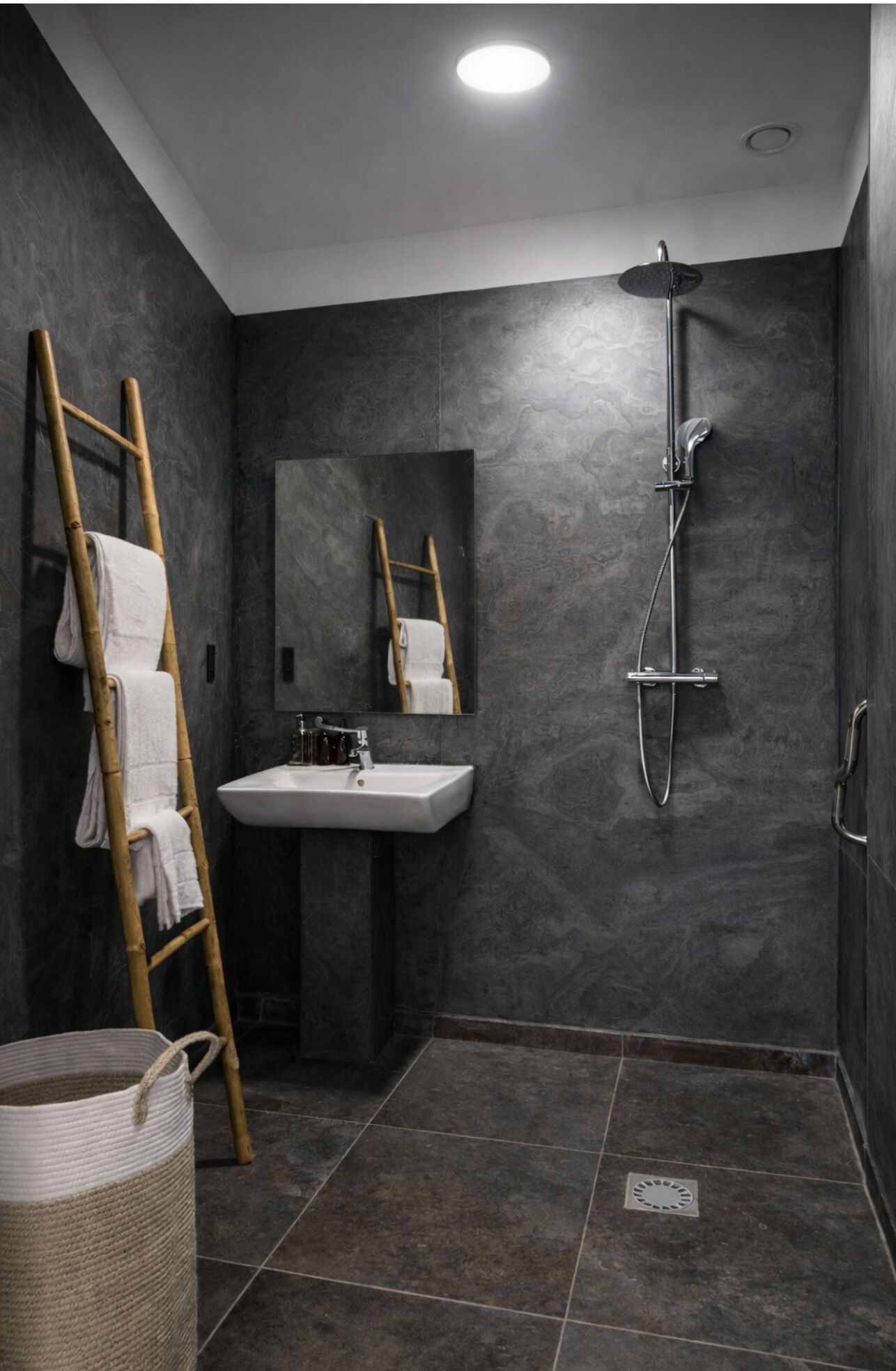 Un baño moderno con paredes de piedra oscura, una escalera de madera con toallas, un lavamanos, un espejo y una ducha.