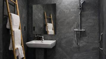 Un baño moderno con paredes de piedra oscura, una escalera de madera con toallas, un lavamanos, un espejo y una ducha.