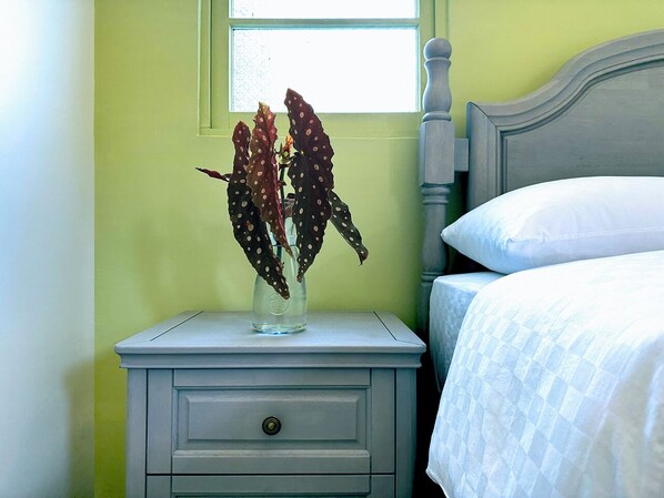 Un dormitorio con una mesita de noche, un florero con flores secas, una cama con ropa de cama blanca y una ventana con luz natural.
