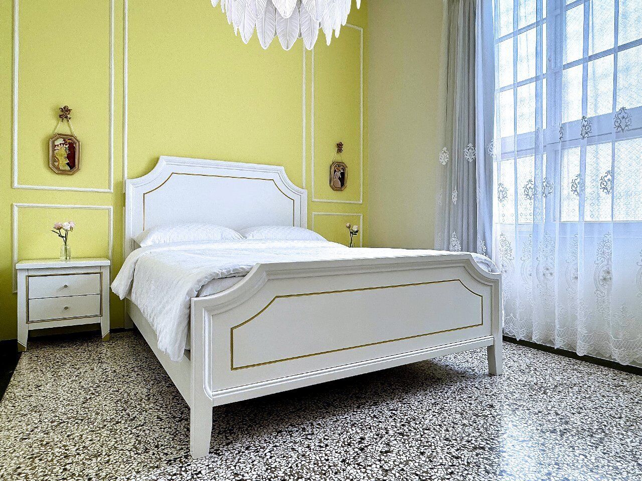 A bedroom with a white bed, a nightstand, a vase with flowers, and a window with curtains.