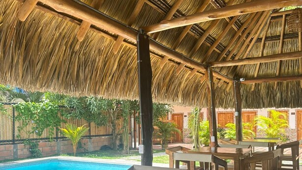 Ein Außenbereich mit Holztischen und -stühlen, einem Reetdach und einem Pool im Hintergrund.