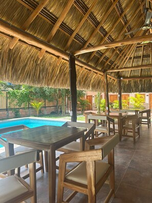 An outdoor seating area with wooden tables and chairs, a thatched roof, and a pool in the background. - Buritaca House (Santa Marta)
