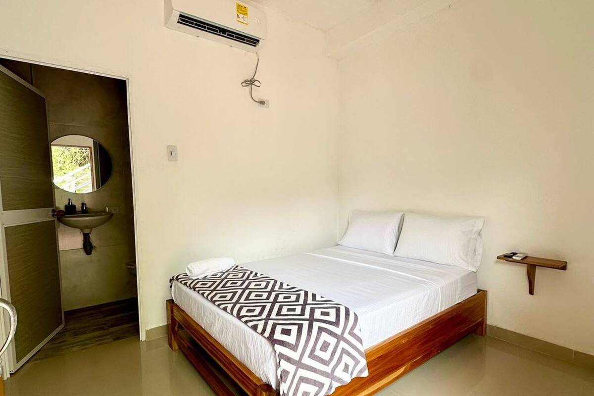 Un dormitorio con una cama de madera, ropa de cama blanca, una mesita de noche pequeña y un aire acondicionado instalado en la pared.