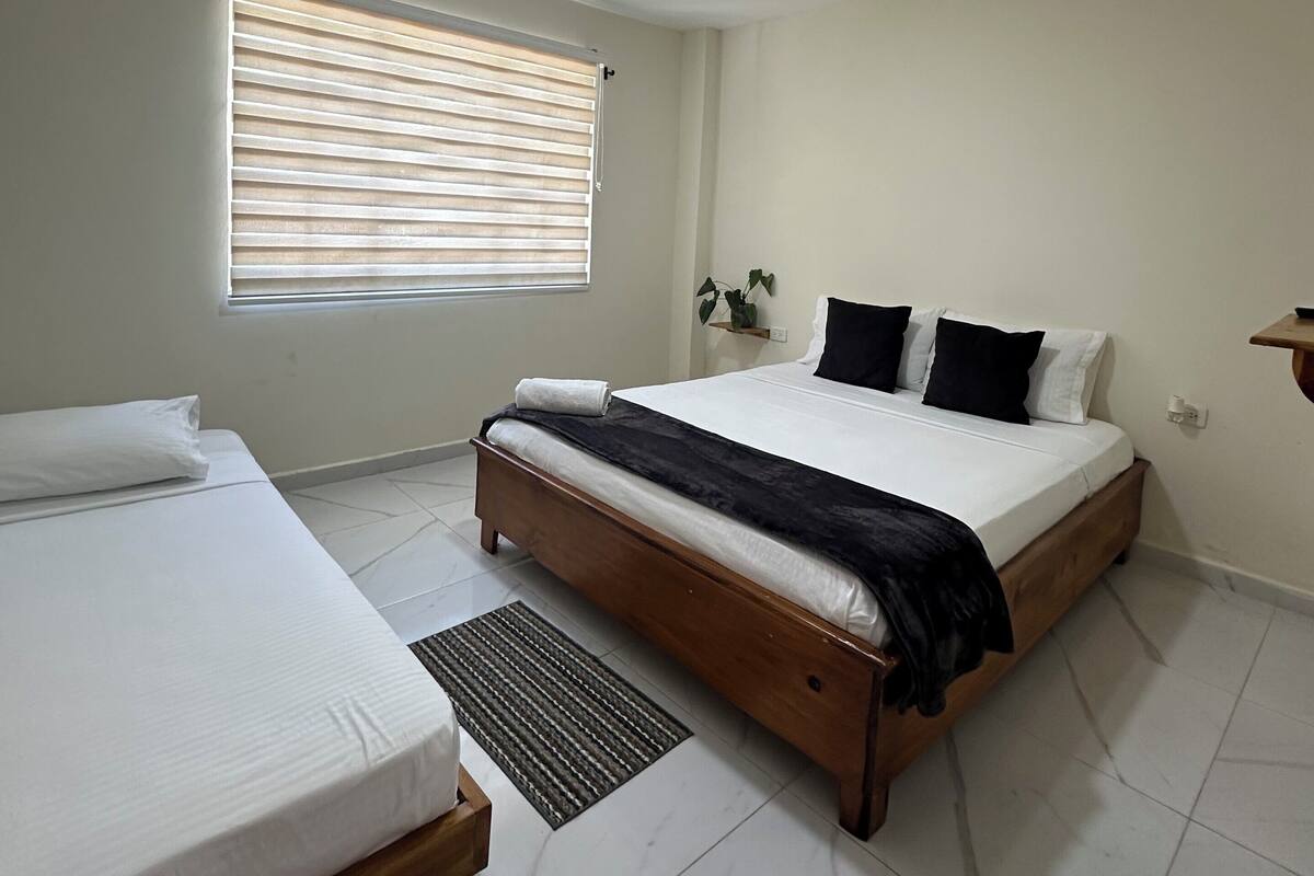 Un dormitorio con una cama grande, una cama más pequeña, una ventana con persianas, una planta y un estante fijado a la pared.