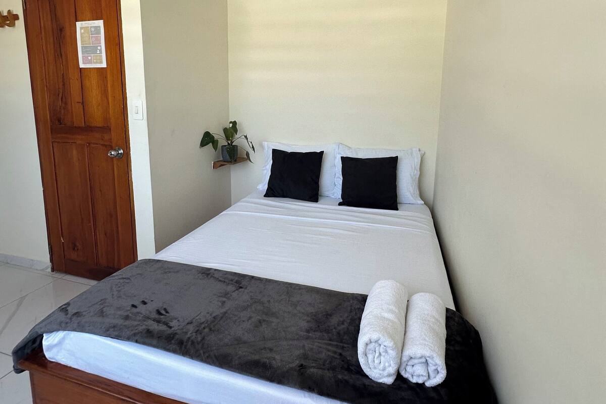 Habitación individual con una cama de madera, ropa de cama blanca y una toalla enrollada sobre la cama.