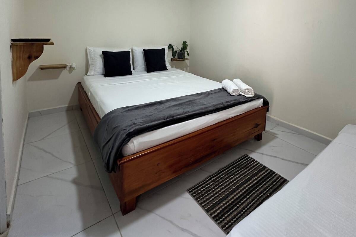 Un dormitorio con una cama de madera, paredes blancas, una lámpara de techo y una mesita de noche pequeña.