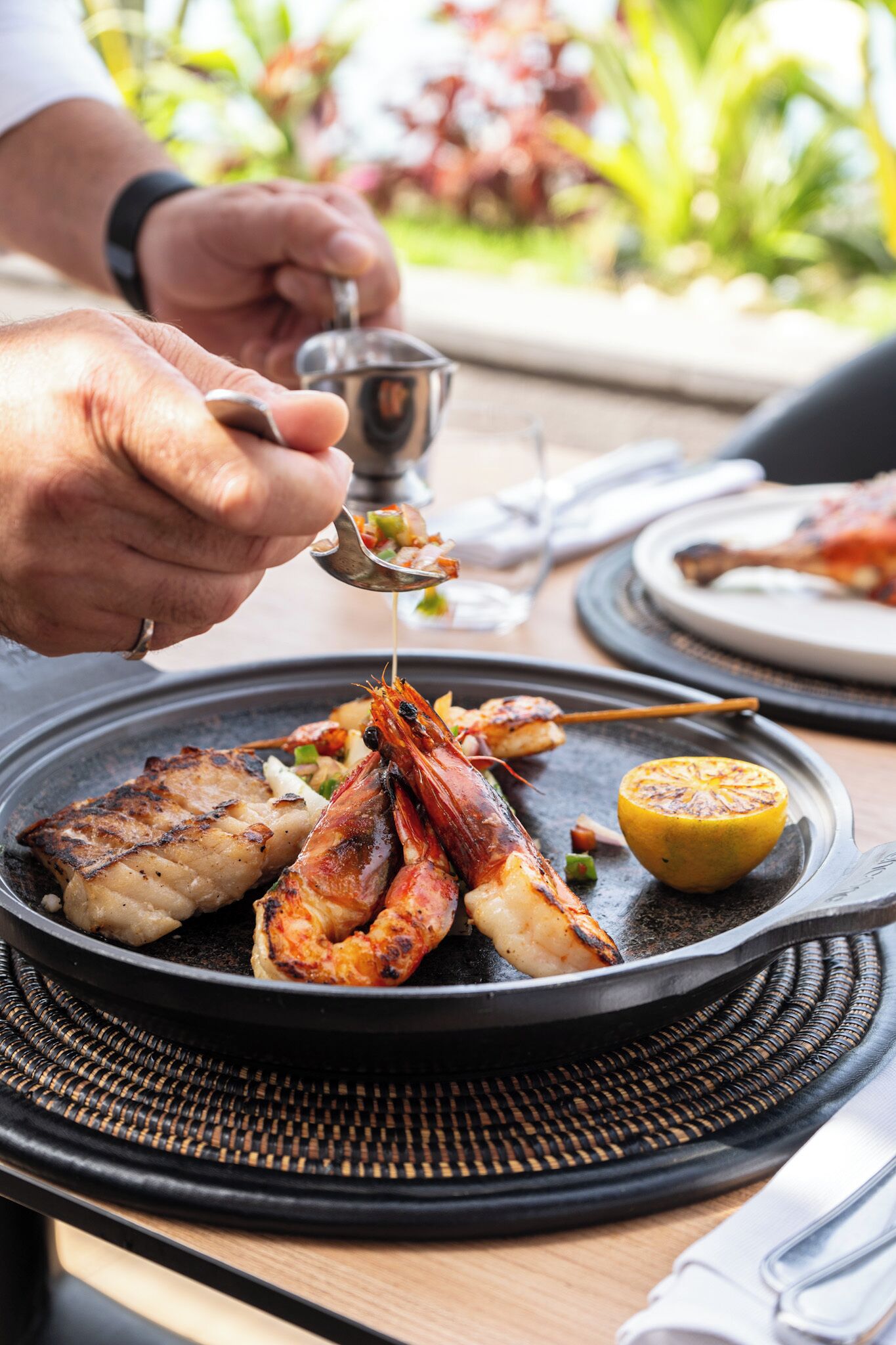 A person serving food from a small metal bowl onto a plate with grilled seafood and a lemon wedge.