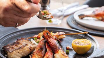 A person serving food from a small metal bowl onto a plate with grilled seafood and a lemon wedge.