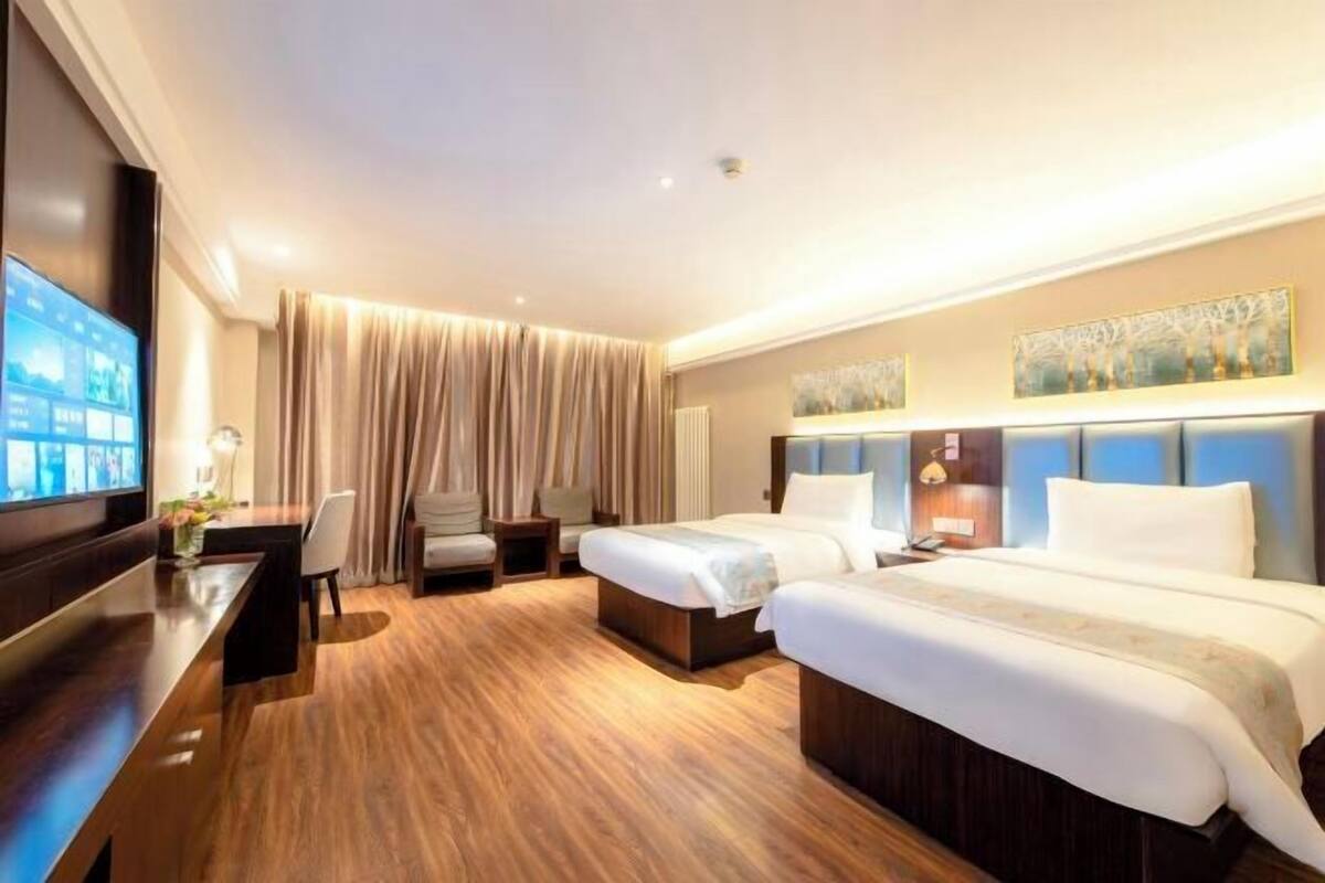 A hotel room with two beds, a desk, a TV, and a large window with curtains.