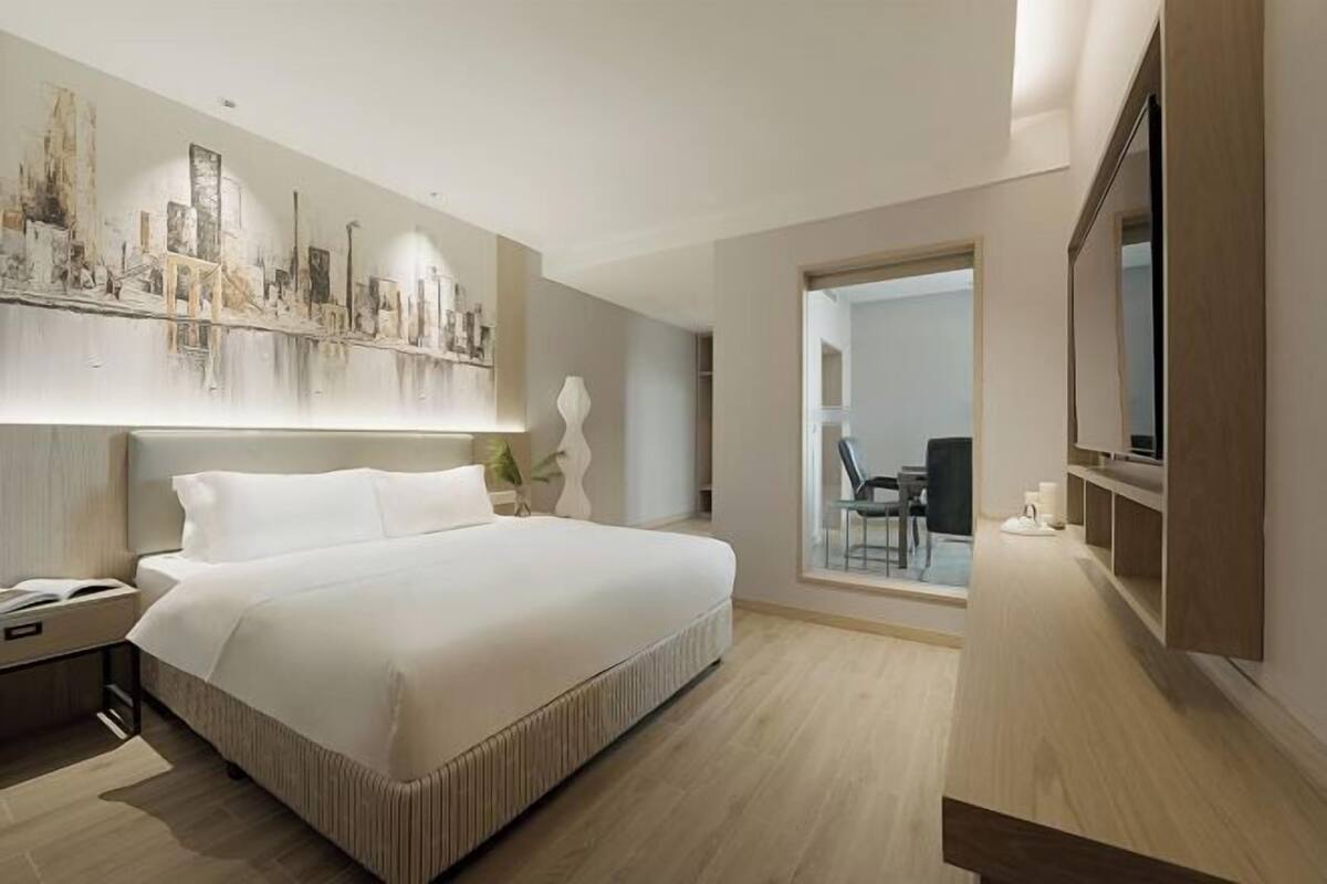 A modern hotel room with a large bed, a cityscape painting on the wall, a desk with a lamp, and a TV on the wall.