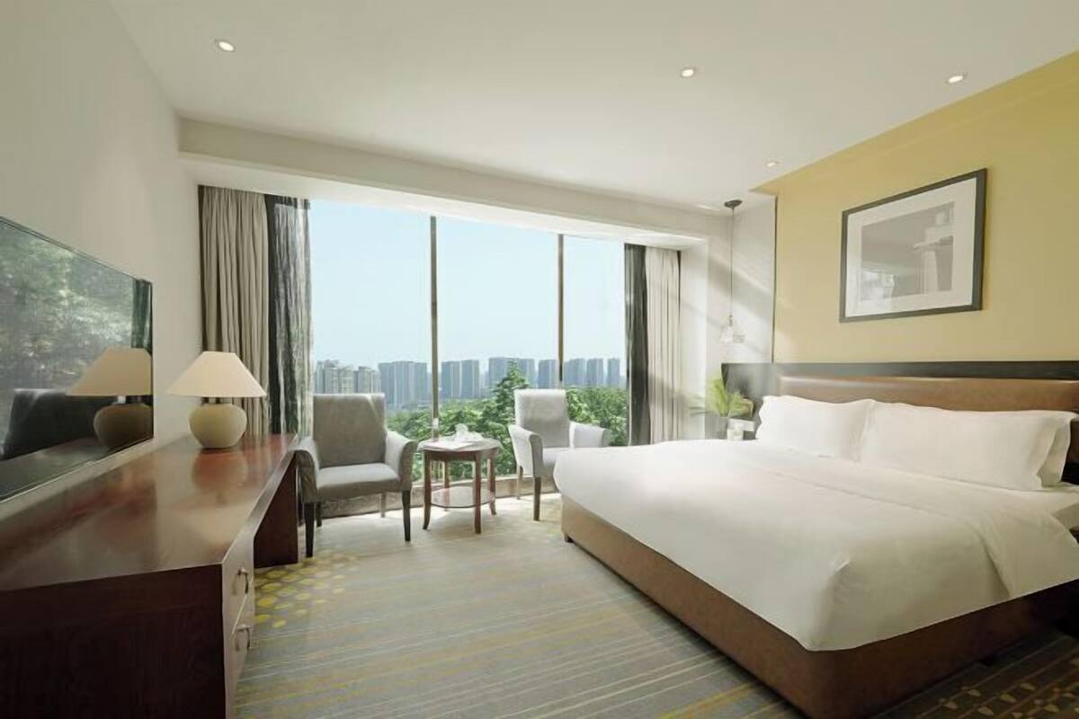 A hotel room with a large bed, a desk with lamps, a chair, a small table, and a large window with a city view.