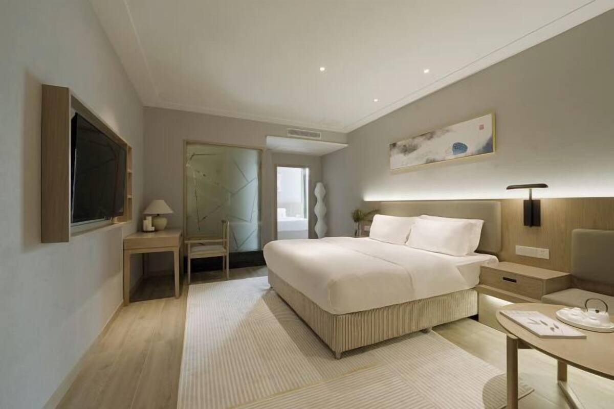 A modern hotel room with a large bed, bedside tables, a desk, and a wall-mounted lamp.