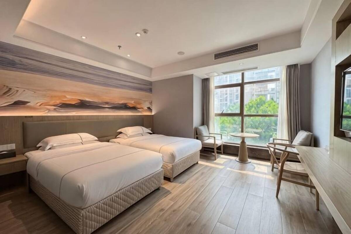 A hotel room with two beds, a dining table, chairs, a large window, and a modern art piece on the wall.