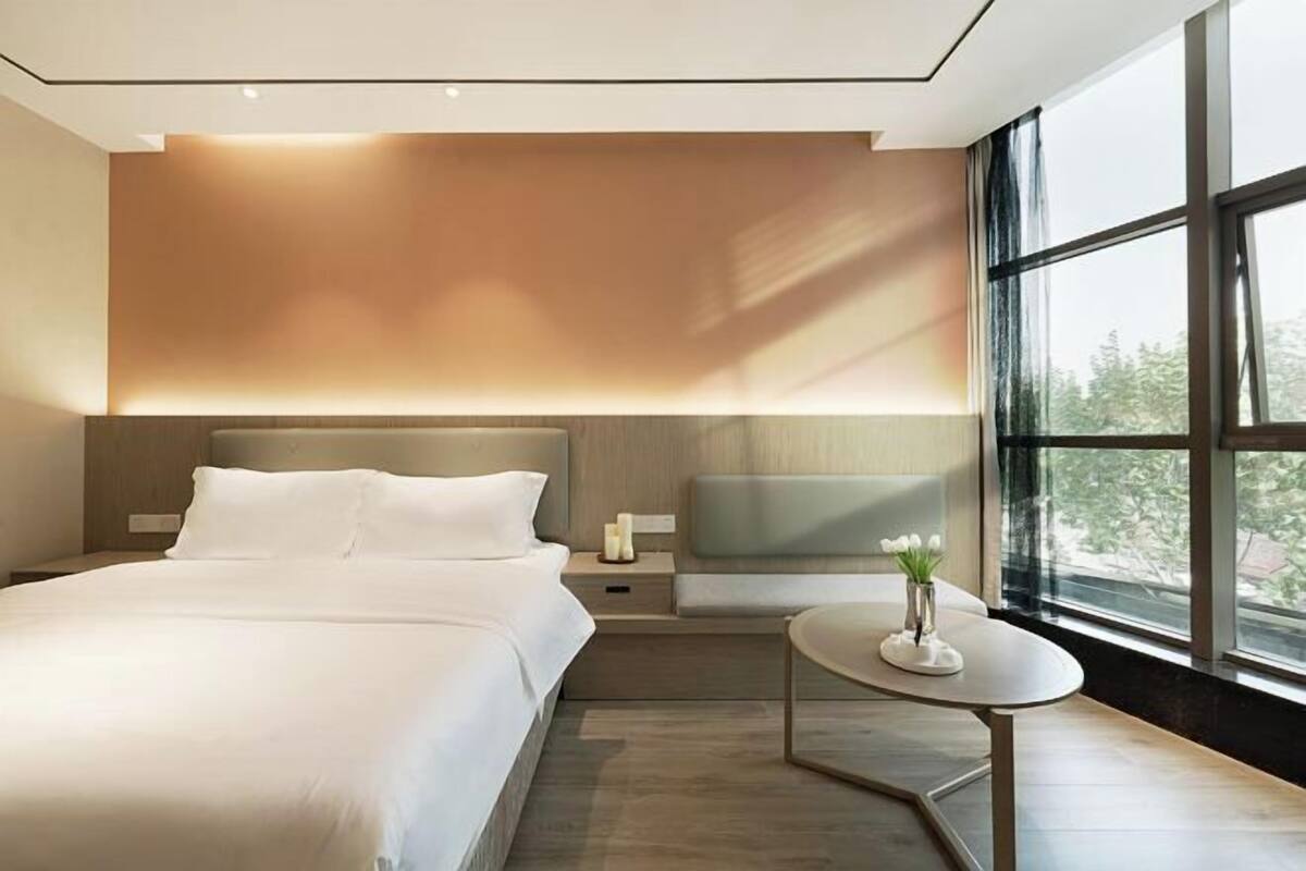 A modern bedroom with a large bed, bedside tables, a small round table with a vase, and a large window providing a view of the outdoors.