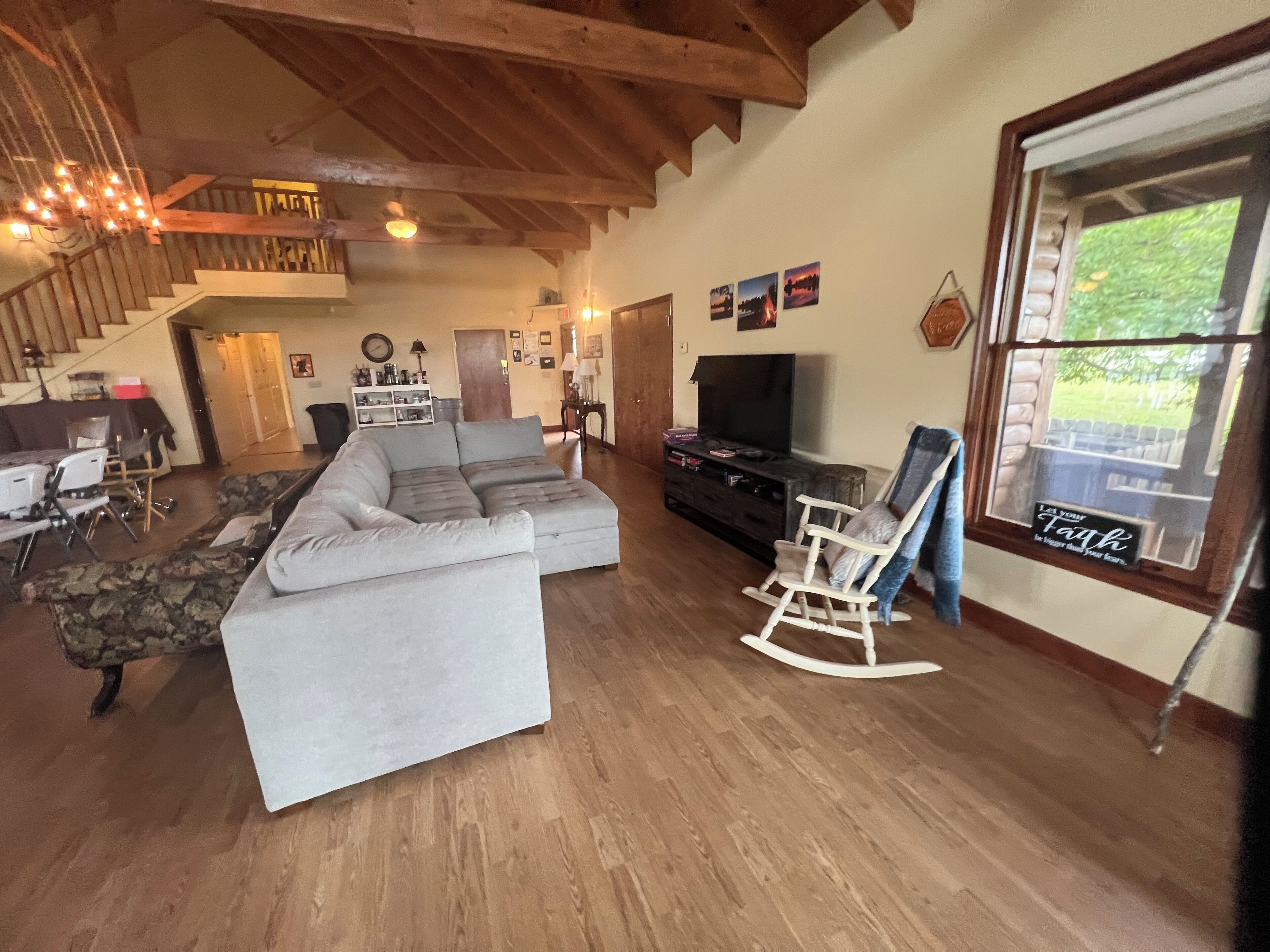 A spacious living room with a large sectional sofa, a television, and a rocking chair. A staircase leads to an upper level, and there is a window with a view of greenery outside.