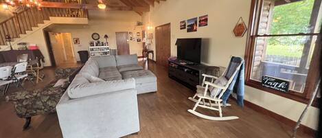 A spacious living room with a large sectional sofa, a television, and a rocking chair. A staircase leads to an upper level, and there is a window with a view of greenery outside.