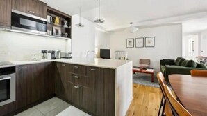 Una cocina moderna con armarios de madera oscura, encimera blanca y electrodomésticos de acero inoxidable. Hay un comedor con una mesa y sillas de madera.