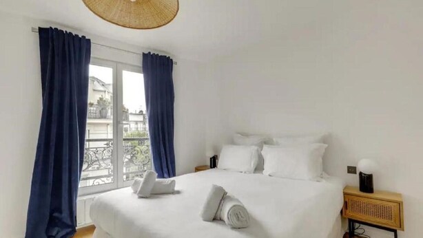 Un dormitorio con cama, ropa de cama blanca, una mesita de noche, una lámpara y vistas a un balcón a través de una ventana con cortinas azules.