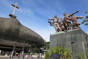A large outdoor sculpture with a cross on top, a building with a curved facade, and people walking around.