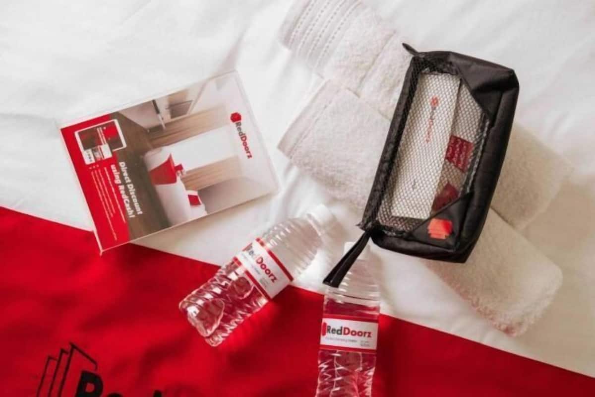 Uma cama com manta branca e vermelha, uma toalha da RedDoorz, uma garrafa de água da RedDoorz e uma caixa da RedDoorz.