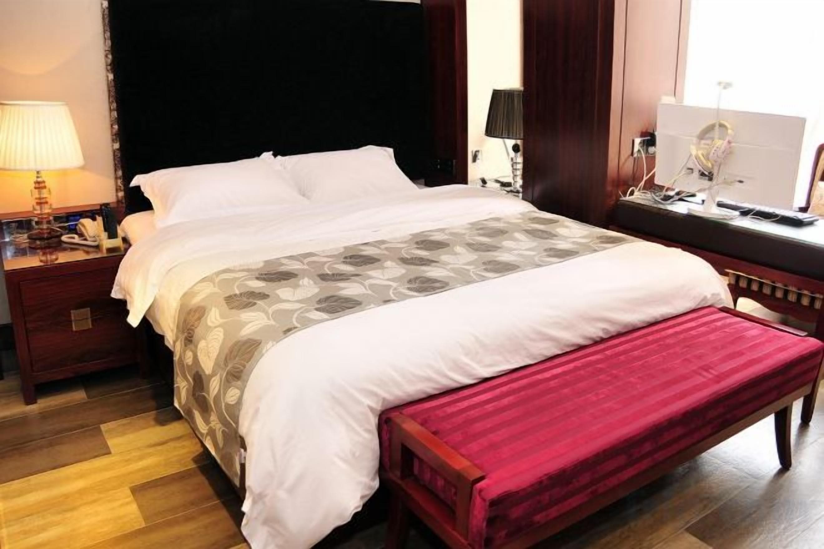 A hotel room with a large bed, a bedside table with a lamp, a red bench, and a desk with a computer.