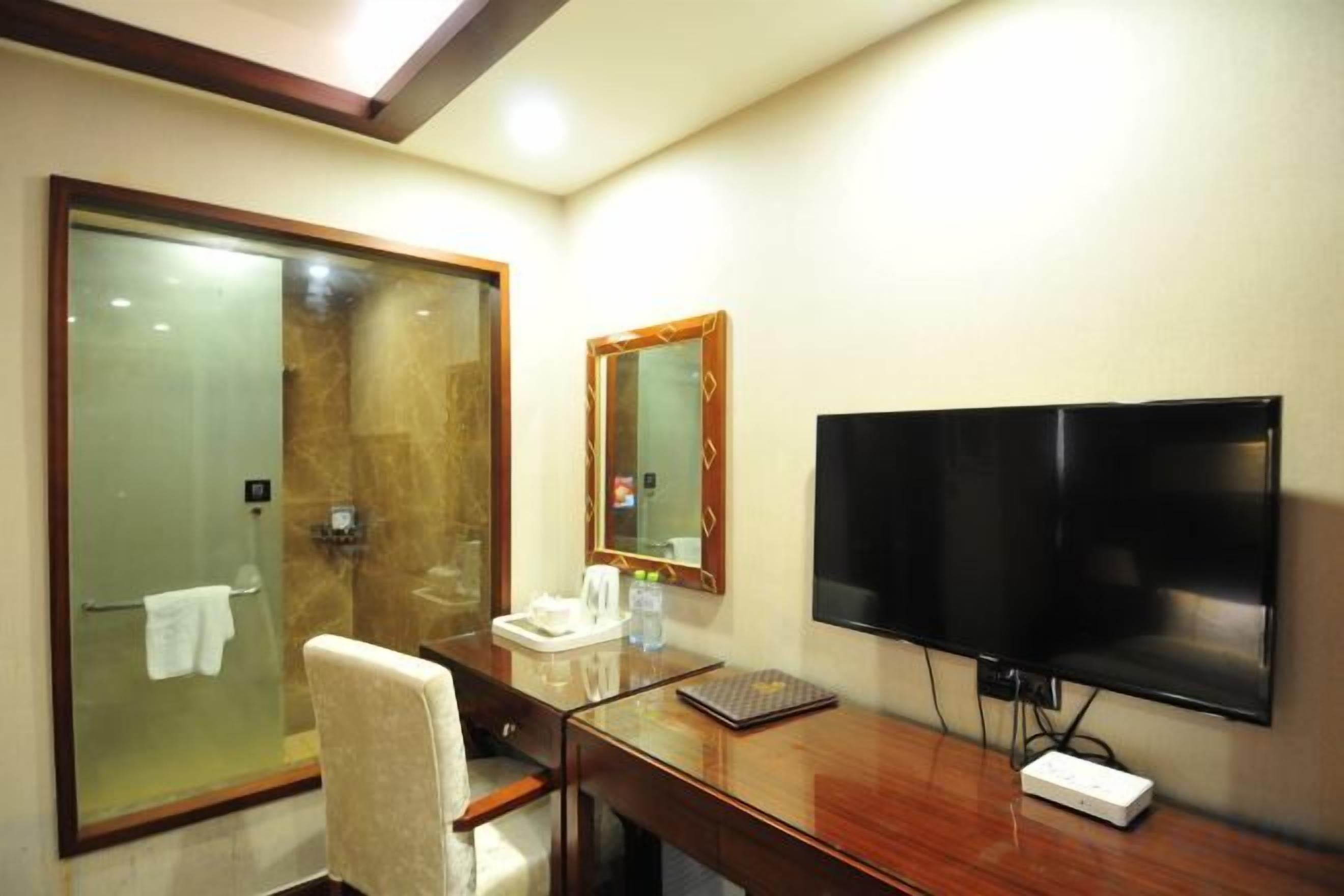 A hotel room with a glass-enclosed shower, a wooden desk with a flat-screen TV, and a mirror.