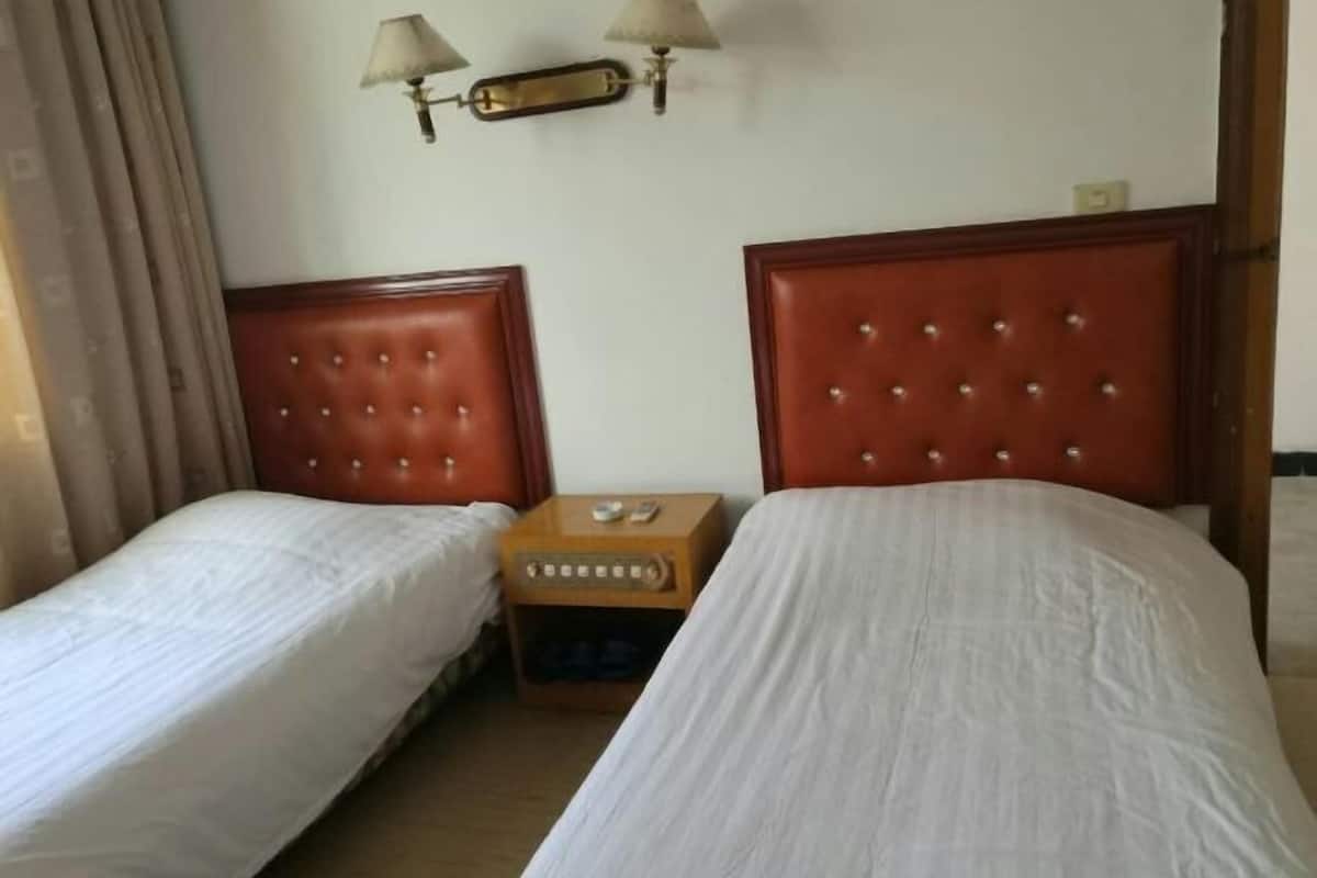 A hotel room with two single beds, a nightstand, a lamp, and a wall-mounted light fixture.