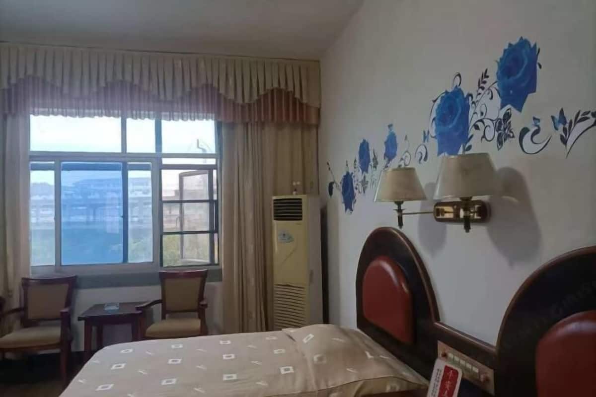 A hotel room with a bed, two chairs, a small table, a window with curtains, a wall-mounted lamp, and a decorative wall with blue floral patterns.