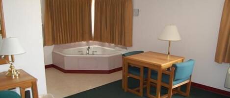 A hotel room with a wooden table and chairs, a small desk with a lamp, a large bathtub, and brown curtains.