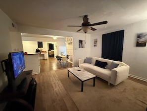 A living room with a sofa, coffee table, TV, ceiling fan, and a view into the kitchen.