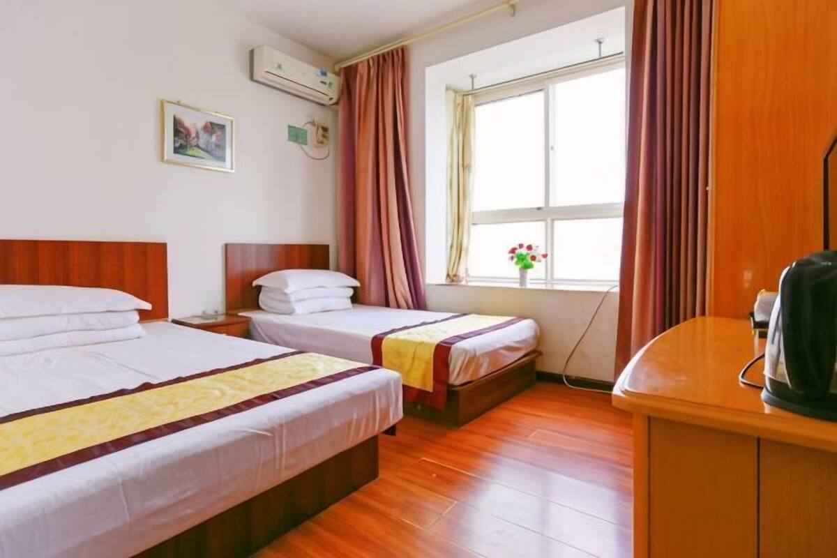Een hotelkamer met twee bedden, een houten vloer, een raam met gordijnen, een wandairconditioner en een kleine tafel met waterkoker.