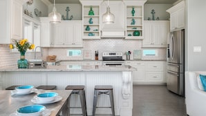 A modern kitchen with white cabinetry, a central island with stools, and a stainless steel refrigerator. - Hot Tub & Direct Beach Access: Walk, Drive or Cart (Bolivar Peninsula)