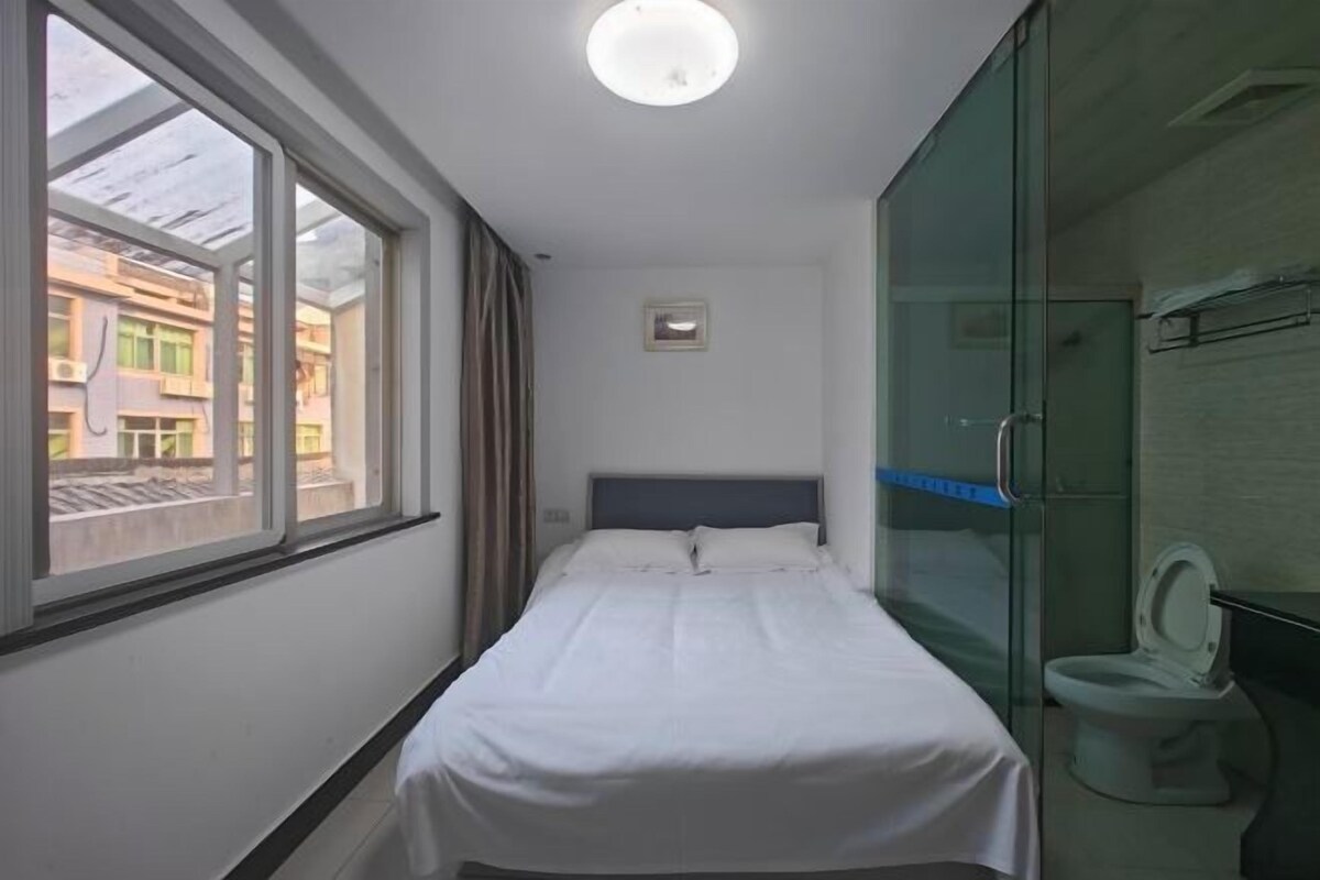 A small hotel room with a single bed, a window, a glass shower enclosure, and a view of buildings outside.