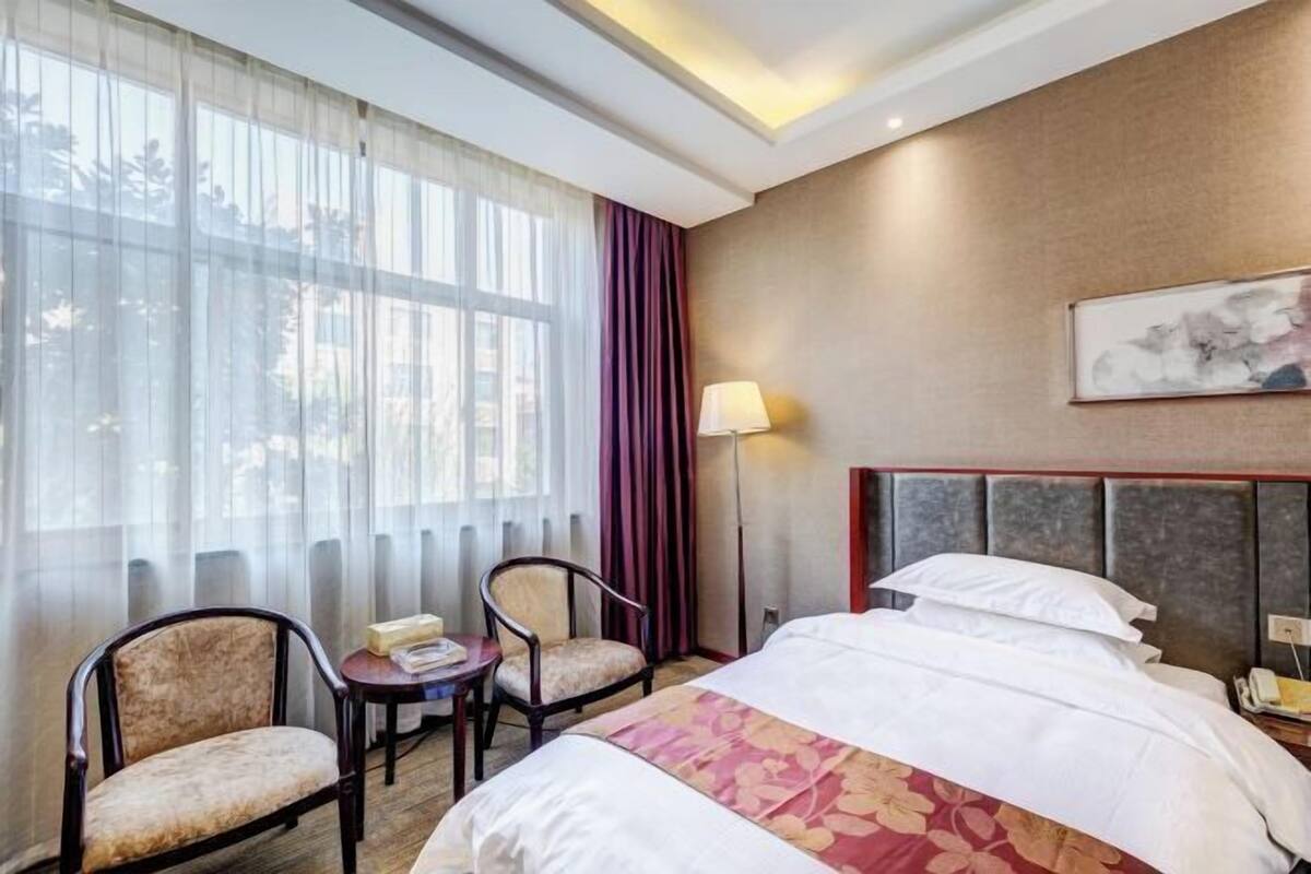 Ein Hotelzimmer mit einem Bett, zwei Stühlen, einem Tisch, einer Lampe und einem Gemälde an der Wand.