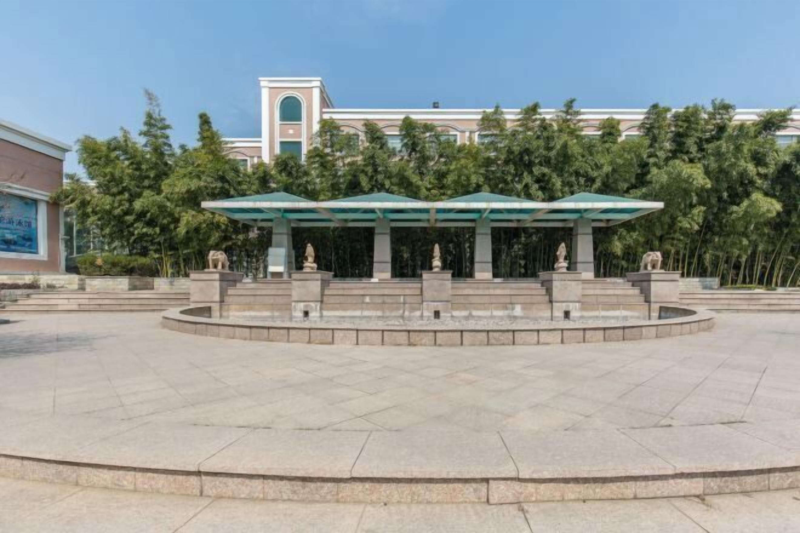 A public square with a central fountain, surrounded by a low stone wall and a covered pavilion with statues.