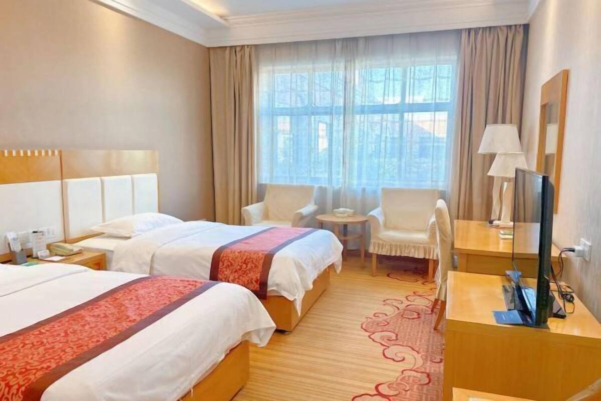 Ein Hotelzimmer mit zwei Betten, einem Schreibtisch, einem Stuhl, einem Fernseher und einem Fenster mit Vorhängen.