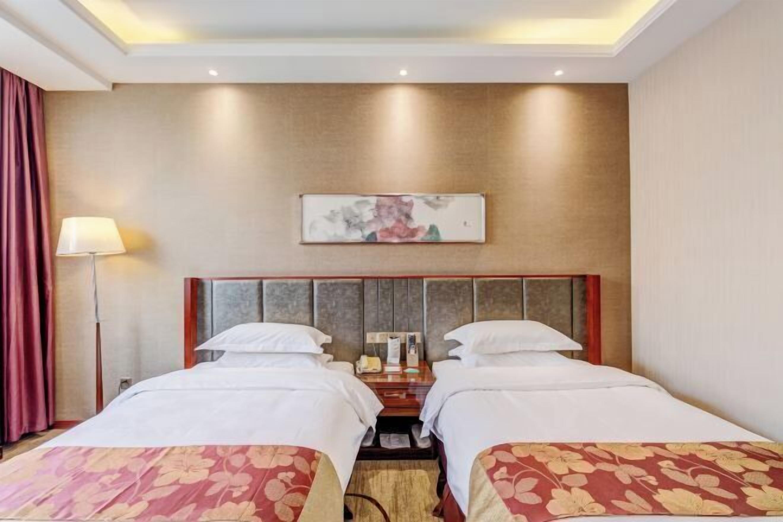 A hotel room with two beds, a headboard, a bedside table, a lamp, and a painting on the wall.