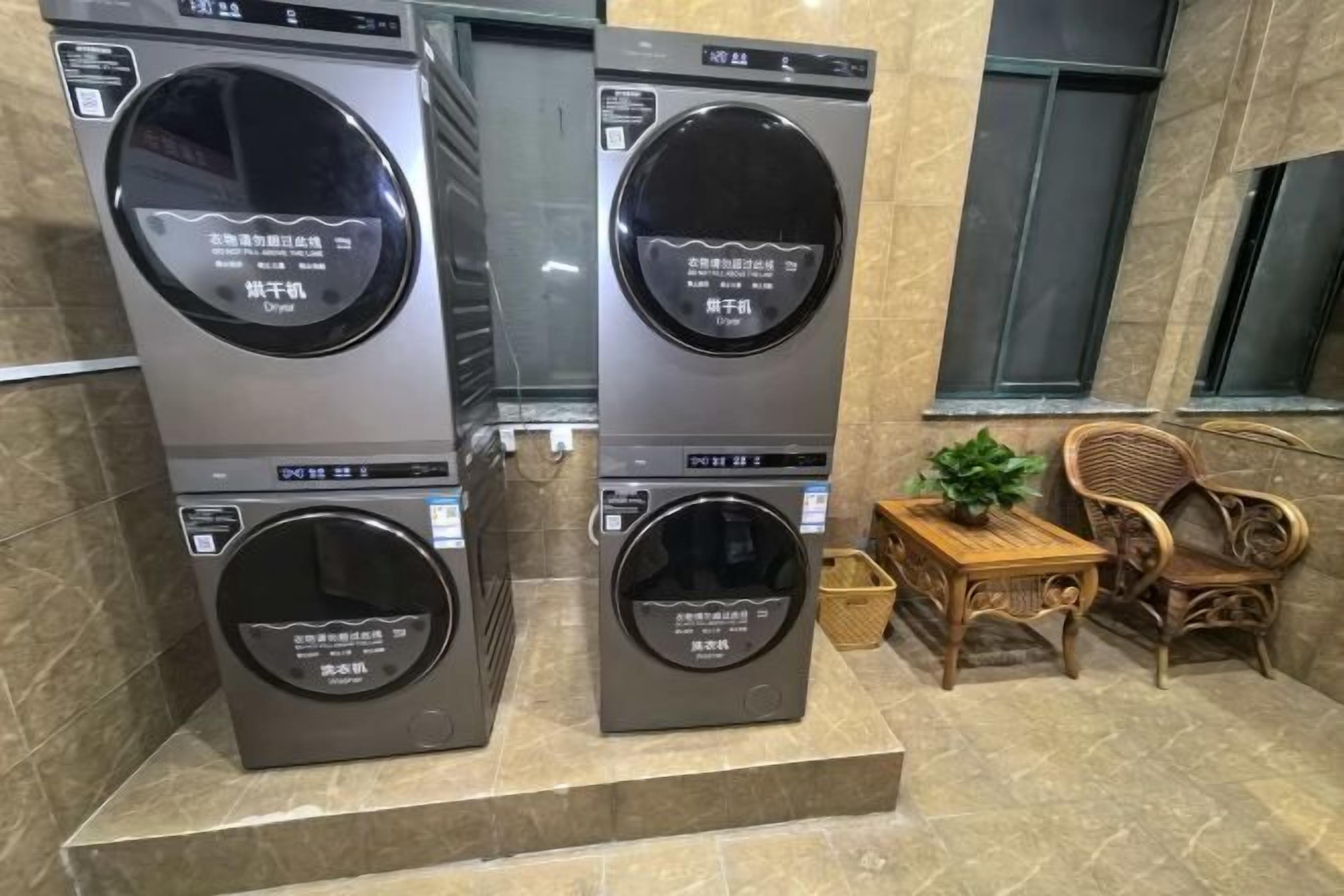 Two stacked washing machines in a public area with a small table and chairs nearby.