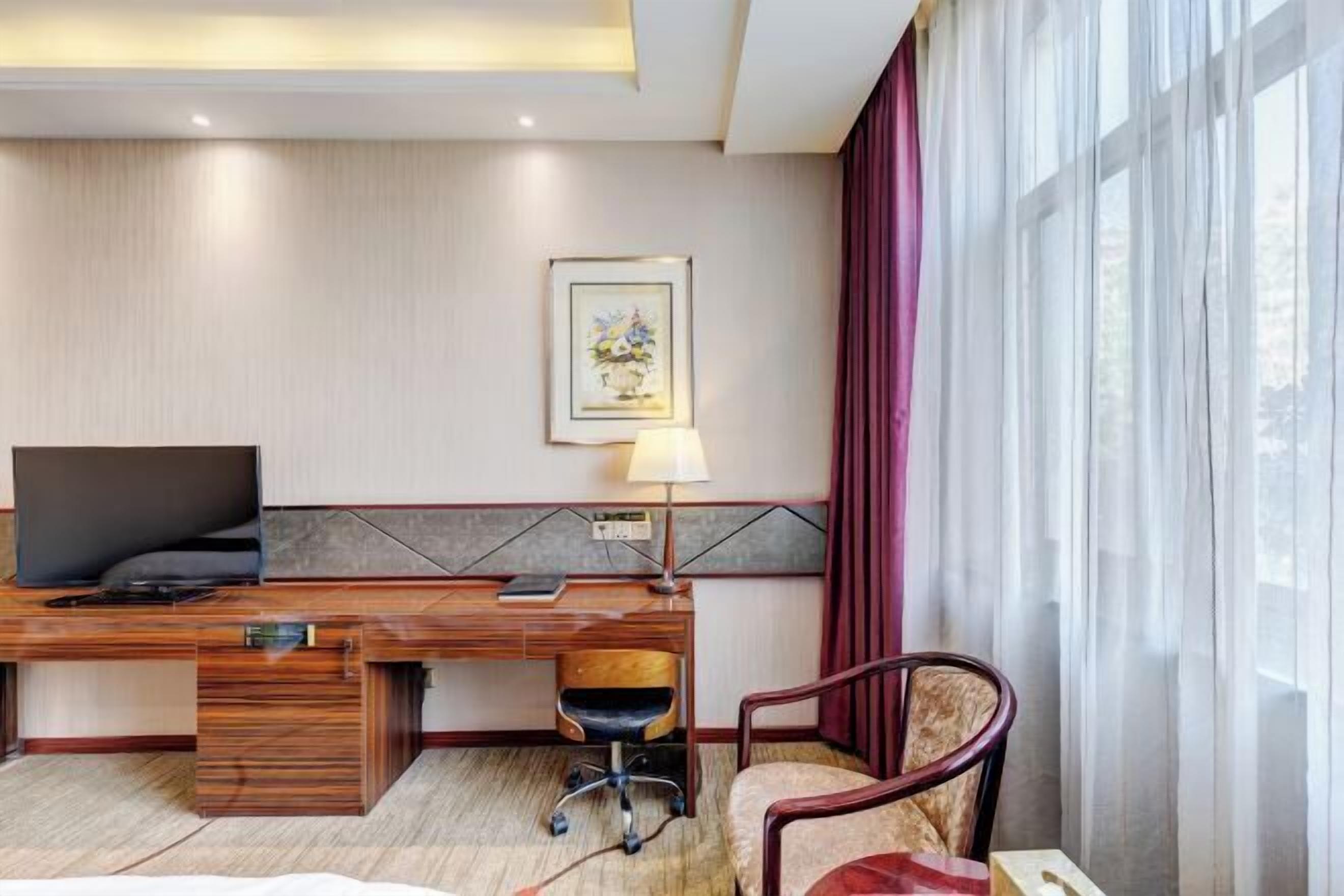 Ett hotellrum med ett skrivbord i trä, en tv, en stol, en lampa och ett fönster med gardiner.