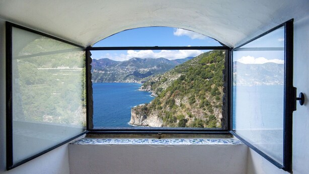 Ein Zimmer mit einem großen Bogenfenster, das einen Blick auf eine Küstenlandschaft mit Bergen und einem Gewässer bietet.