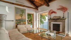 Ein geräumiges Wohnzimmer mit einem Glastisch, beigefarbenen Sofas, einem Kamin und Blick aufs Meer.