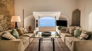 Ein Wohnzimmer mit einem großen Bogenfenster und Meerblick, zwei Sofas mit gemusterten Kissen, einem Glastisch und einer Stehlampe.
